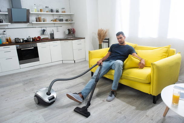 Young man holding vacuum cleaner while sitting on couch at home  - Valokuva, kuva