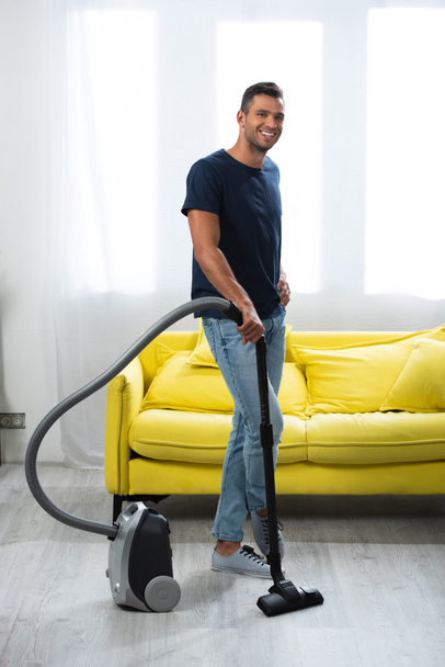 Smiling man with hand on hip holding brush of vacuum cleaner at home  - Fotografie, Obrázek