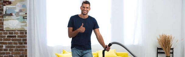 Glimlachende man toont als gebaar terwijl het houden van borstel van stofzuiger thuis, banner  - Foto, afbeelding