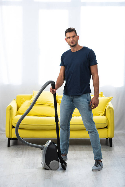 Young man holding vacuum cleaner near sofa in living room  - Фото, изображение