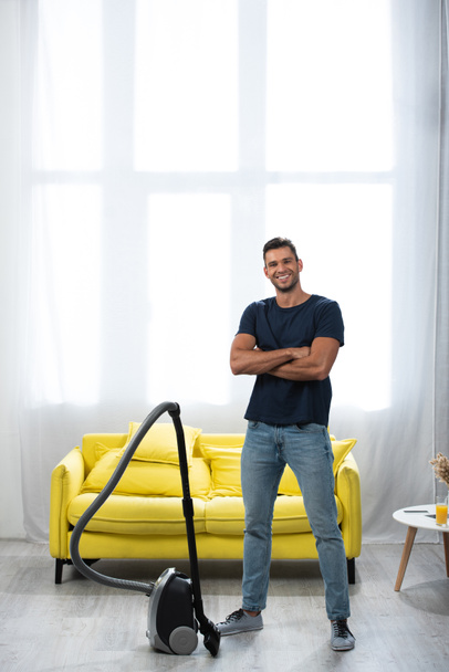 Cheerful man with crossed arms standing near vacuum cleaner at home  - Photo, Image