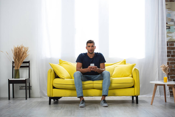 Excited man holding smartphone while looking at camera on couch  - Фото, изображение