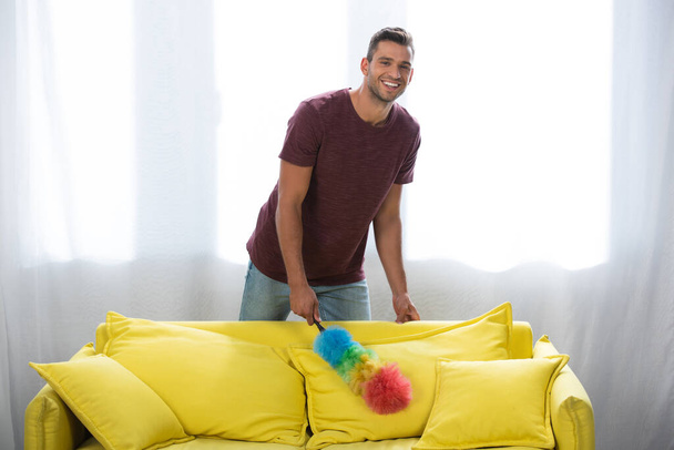 Homme souriant avec brosse à poussière debout près du canapé jaune à la maison  - Photo, image