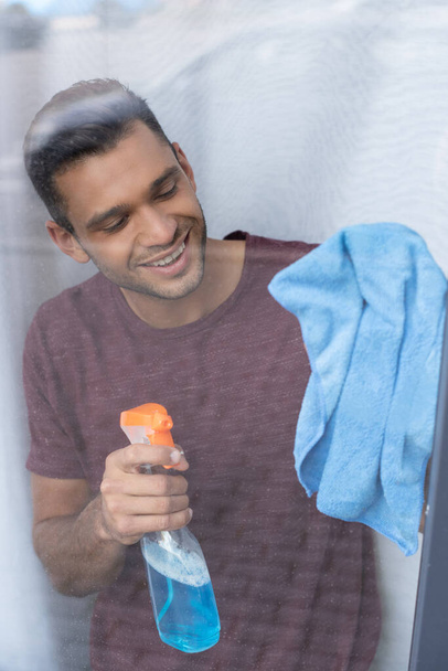 Glimlachende man schoonmaken van glas van het raam met wasmiddel en doek  - Foto, afbeelding