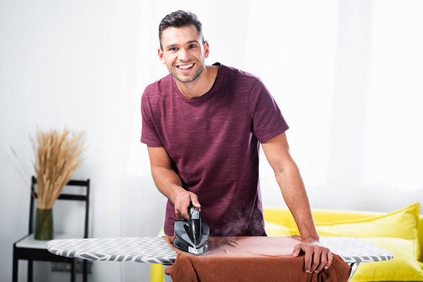 Cheerful man using iron while ironing clothes on board in living room  - Foto, imagen