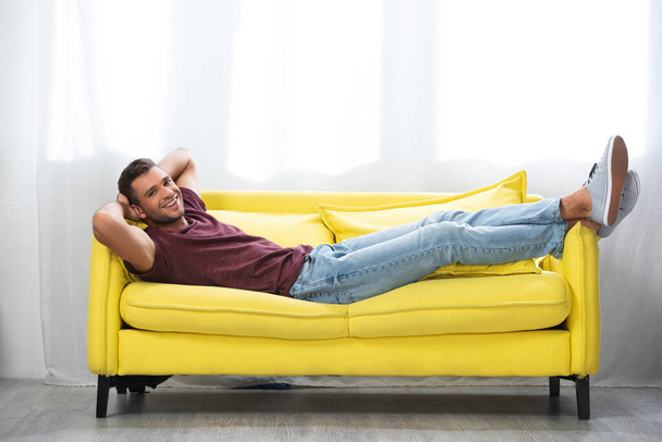 Allegro uomo guardando la fotocamera mentre sdraiato con le gambe incrociate sul divano  - Foto, immagini