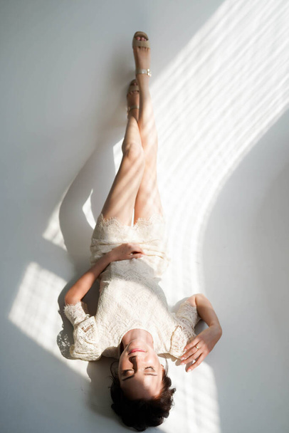 Softly sunlights on young brunette woman in white dress lying on white floor in studio - 写真・画像