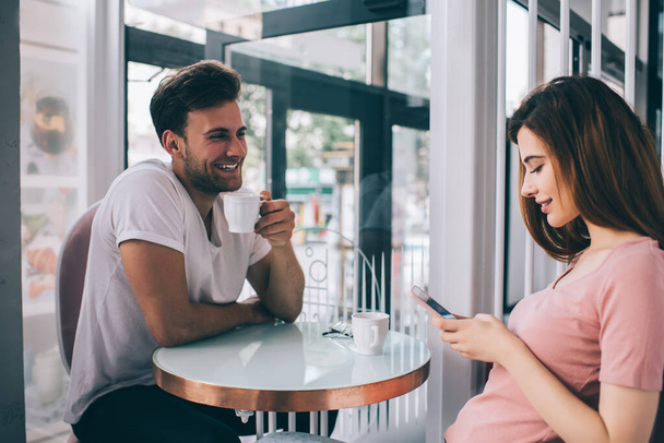 Happy young handsome man drinking cup of coffee and looking at charming woman using smartphone while sitting together at table in cafeteria - Photo, image