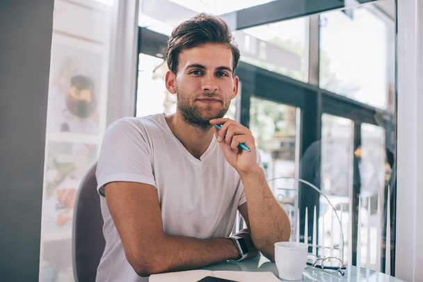 Satisfied youthful handsome unshaven male in casual clothes looking at camera and touching chin with pen while having break and sitting at table in cafeteria - Foto, Bild