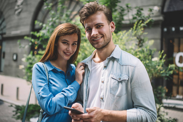 Pleased adult man in casual clothes and delighted woman looking at camera while standing on street and using smartphone against blurred old building and green trees in daytime - Foto, immagini