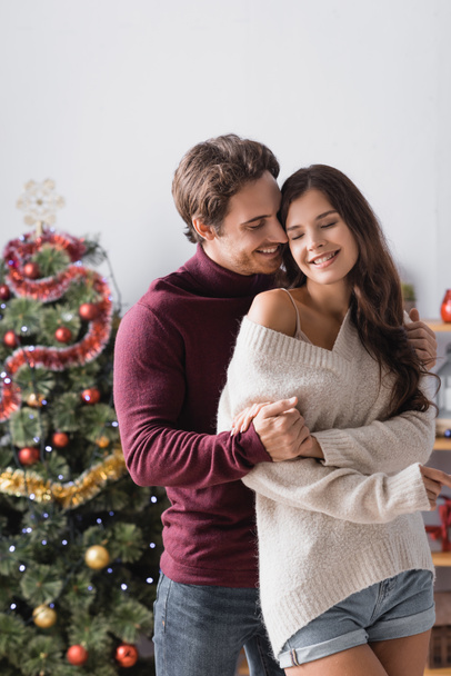 pleased couple in warm sweaters hugging near decorated christmas tree on blurred background - Foto, Bild