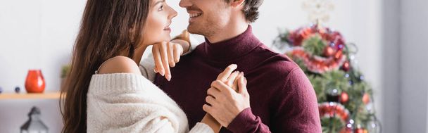 joyful couple in sweaters holding hands near decorated christmas tree on blurred background, blur - Photo, Image