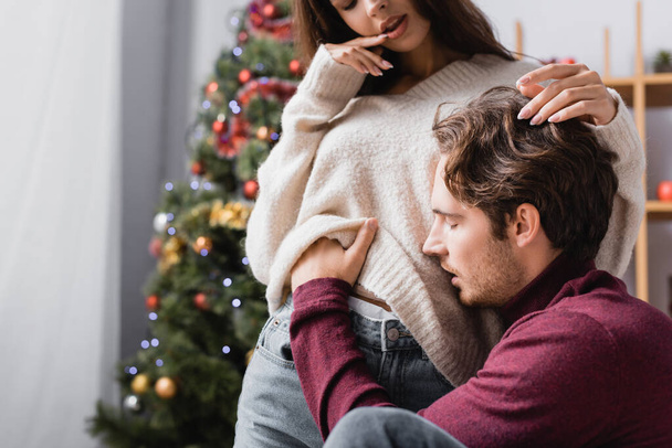 passionate man undressing young woman in sweater near christmas tree on blurred background - Photo, Image