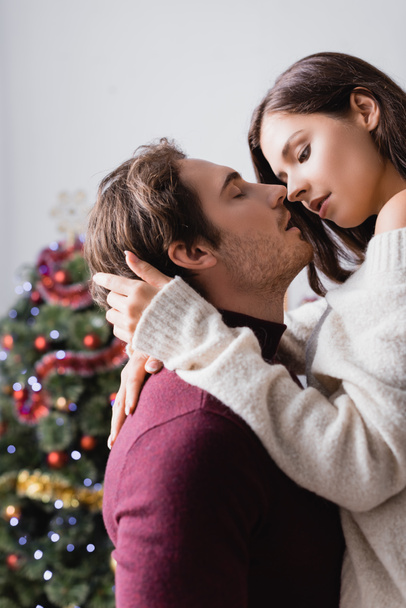 passionate man embracing girlfriend in sweater near christmas tree on blurred background - 写真・画像