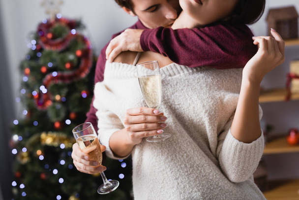 vue recadrée de l'homme passionné embrasser femme tout en tenant des verres avec du champagne près de l'arbre de Noël sur fond flou - Photo, image