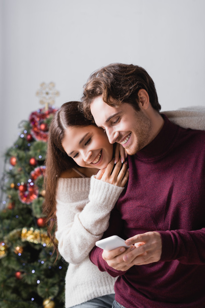 幸せなカップルのセーターを着てクリスマスツリーの近くのスマートフォンを見て背景にぼやけて - 写真・画像