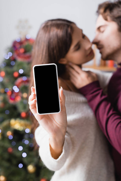 Frau hält Smartphone mit leerem Bildschirm neben Freund und geschmücktem Weihnachtsbaum auf verschwommenem Hintergrund - Foto, Bild