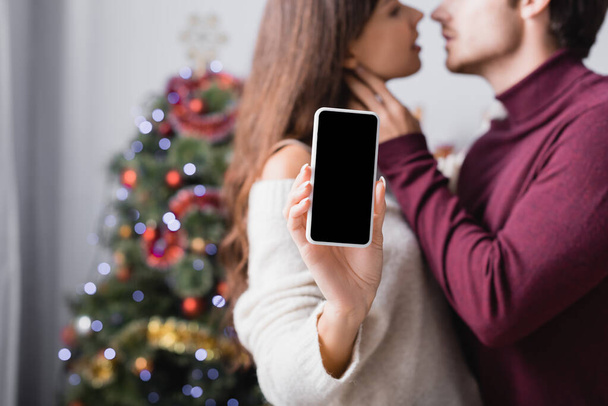 mujer sosteniendo teléfono inteligente con pantalla en blanco cerca de novio y árbol de Navidad sobre fondo borroso - Foto, imagen