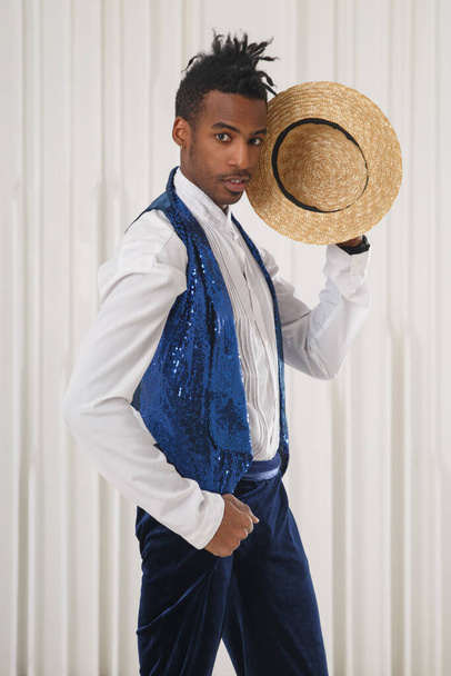 Un giovane ragazzo nero in abiti da palcoscenico tiene un cappello di paglia vicino al suo viso su uno sfondo leggero. - Foto, immagini