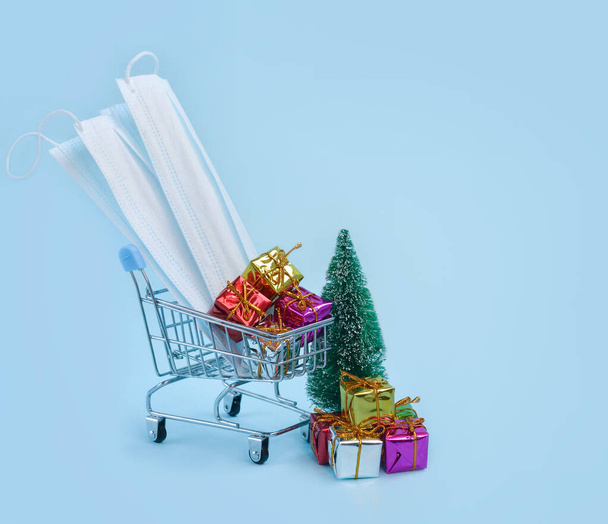 ギフトボックス、クリスマスツリー、医療用マスク、青い背景にショッピングトロリー隔離中の安全なオンラインショッピングとコロナウイルスの普及の概念。スペースフットテキストのコピー. - 写真・画像