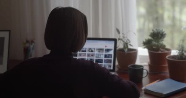 Pohled zezadu na dívku sedící u stolu se zápisníkem pracujícím doma zpomaleně. Nerozpoznatelná žena na volné noze hledající fotografie na notebooku přirozeného světla. Microstock freelance business - Záběry, video