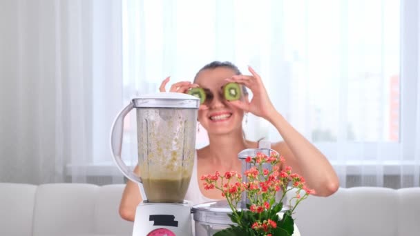 feliz joven mujer preparando delicioso batido nutritivo en licuadora. divertida hembra haciendo ojos desde kiwi. - Imágenes, Vídeo