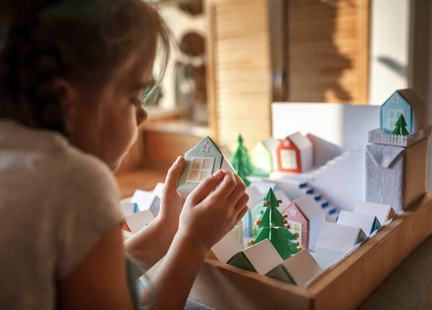 Origami calendario de adviento, pueblo de Navidad, artesanía de papel. Linda chica mirando pequeñas casas de papel con el número y el árbol verde de papel, actividad estacional con los niños, estilo de vida interior - Foto, Imagen