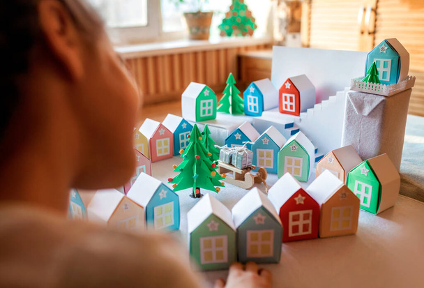 Origami advent calendar, Christmas village, paper craft. Cute girl looking upon tiny paper houses with number and paper green tree, seasonal activity with kids, indoor lifestyle - Photo, Image