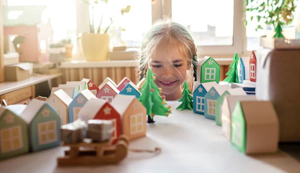 Origami adventskalender, Kerst dorp, papier ambachtelijke. Schattig meisje op zoek naar kleine papieren huizen met nummer en papier groene boom, seizoensgebonden activiteit met kinderen, indoor lifestyle - Foto, afbeelding