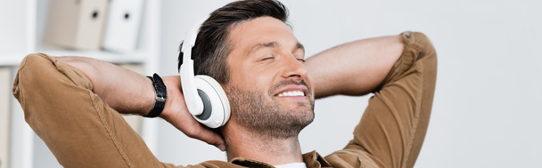 Hombre de negocios sonriente con auriculares, con las manos detrás de la cabeza y escuchando música sobre fondo borroso, pancarta - Foto, imagen