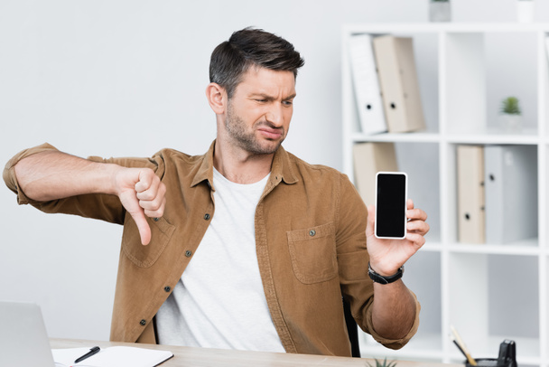 Dissatisfied businessman with thumb down, holding smartphone with blank screen at workplace on blurred background - Photo, Image