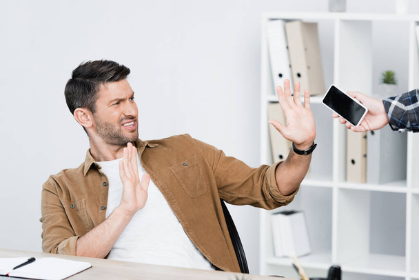Podenerwowany biznesmen z zatrzymaniem gestu patrząc na smartfona w ręku człowieka w pobliżu miejsca pracy - Zdjęcie, obraz