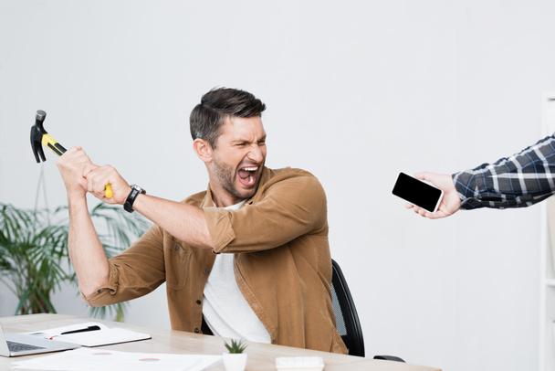 Aggressiivinen liikemies vasaralla huutaa istuessaan lähellä älypuhelinta kädessä työpaikan lähellä - Valokuva, kuva
