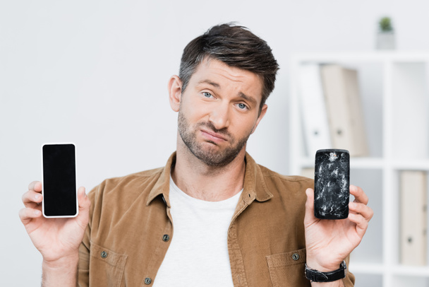 Niezadowolony biznesmen patrząc w kamerę trzymając rozbity smartfon i komórkę z pustym ekranem - Zdjęcie, obraz