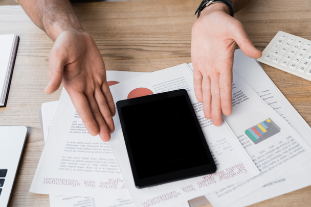 Ausgeschnittene Ansicht eines Geschäftsmannes, der mit den Händen auf ein kaputtes digitales Tablet auf strukturiertem Holzhintergrund mit Papierblättern zeigt - Foto, Bild