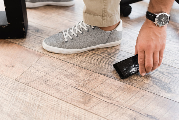 Vista ritagliata di uomo d'affari che prende smartphone danneggiato dal pavimento in legno - Foto, immagini