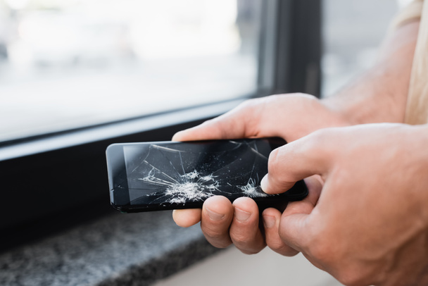 Cropped view of businessman touching smashed touchscreen of smartphone with big finger on blurred background - Photo, Image