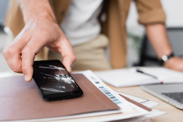 Обрізаний вид бізнесмена, який бере смартфон з тріщинами з купи паперових робіт на розмитому фоні
 - Фото, зображення