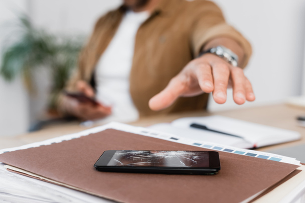 Обрезанный вид руки бизнесмена рядом с разбитым смартфоном на куче бумажной работы на рабочем месте на размытом фоне - Фото, изображение