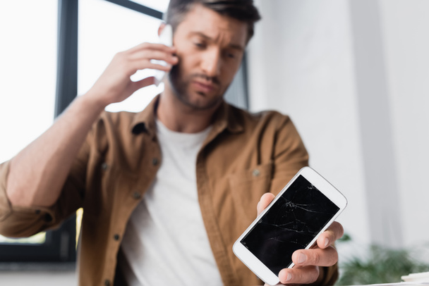 Zertrümmertes Smartphone in der Hand eines Geschäftsmannes, der auf dem Handy mit verschwommenem Fenster im Hintergrund spricht - Foto, Bild