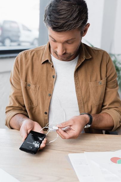 Empresario con auriculares, sosteniendo el teléfono inteligente roto mientras está sentado en el lugar de trabajo sobre un fondo borroso - Foto, Imagen
