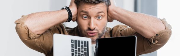 Homme d'affaires fou avec les mains près de la tête regardant ordinateur portable cassé sur fond flou, bannière - Photo, image
