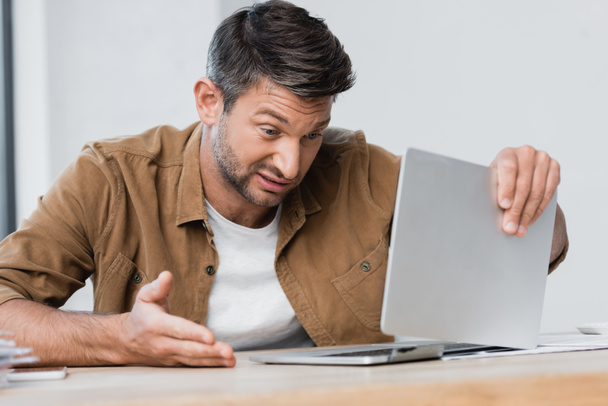 Незадоволений бізнесмен жестикулює, дивлячись на зламаний ноутбук на робочому місці на розмитому передньому плані
 - Фото, зображення