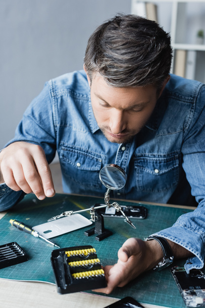 Repairman looking at holder with screwdriver bits at workplace with disassembled mobile phones on blurred background - Fotografie, Obrázek