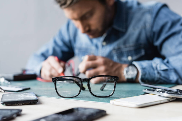 Szemüveg az asztalon közelében szétszerelt alkatrészek mobiltelefonok elmosódott szerelő dolgozik a háttérben - Fotó, kép