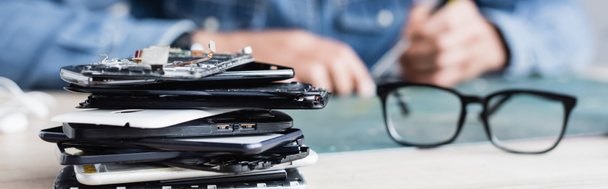 Close up view of pile of broken mobile phones near eyeglasses on workplace with blurred repairman on background, banner - Фото, изображение