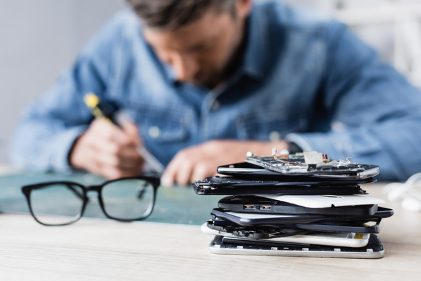 Close up view of pile of broken mobile phones near eyeglasses on workplace with blurred repairman on background - Photo, Image