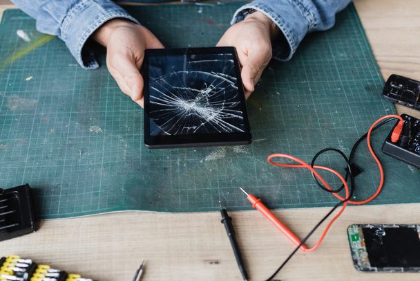 Обрезанный вид ремонтника с разбитым цифровым планшетом на рабочем месте возле мультиметра и разобранными мобильными телефонами - Фото, изображение