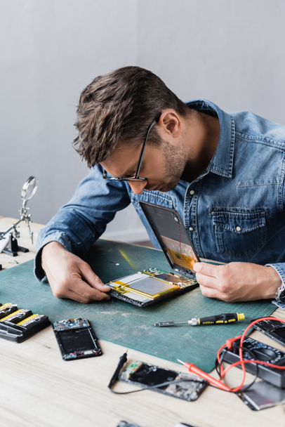 Repairman in eyeglasses looking at disassembled broken digital tablet near screwdriver and multimeter at workplace - Photo, Image