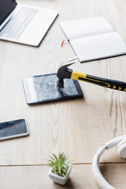 Hamer op verbrijzelde digitale tablet in de buurt van koptelefoon en blanco notebook op tafel met digitale apparaten - Foto, afbeelding